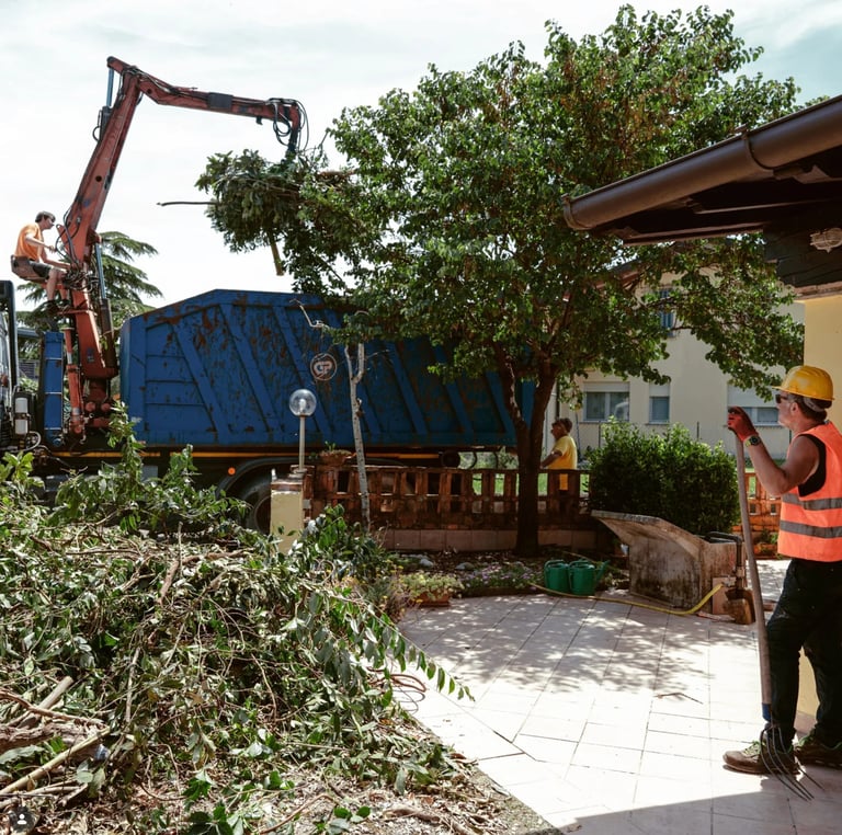 A worker operates a red hydraulic arm to load green plant material into a blue waste bin at an outdoor facility